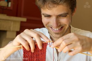 Close up image of smiling man knitting with timber needles and red wool. Read our blog. Healing stitches: The therapeutic benefits of knitting and crocheting.